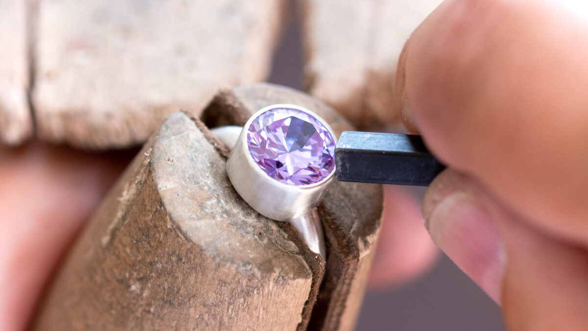 A large round faceted lavender cubic zirconia being set in a silver ring with a pusher tool