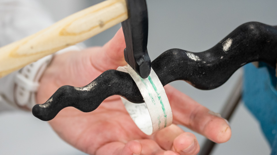 a silver bangle being hammered on an anticlastic raising stake against a plain grey background