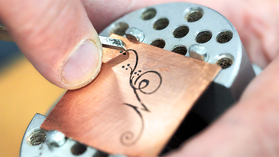 A copper sheet with a hand drawn calligraphic letter N being engraved using a graver