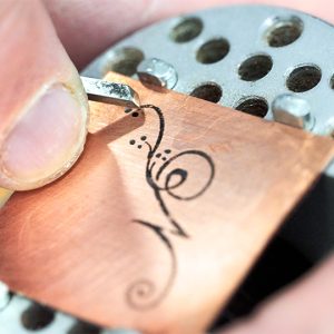A copper sheet with a hand drawn calligraphic letter N being engraved using a graver