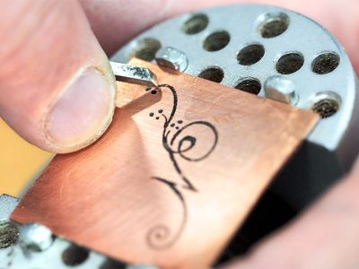 A copper sheet with a hand drawn calligraphic letter N being engraved using a graver