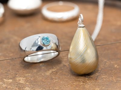 Hollow form silver jewellery on a jewellery bench including rings and a teardrop shaped textured pendant with gold embellishment.