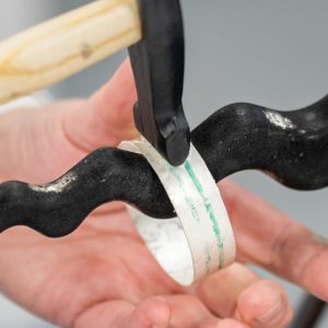 a silver bangle being hammered on an anticlastic raising stake against a plain grey background